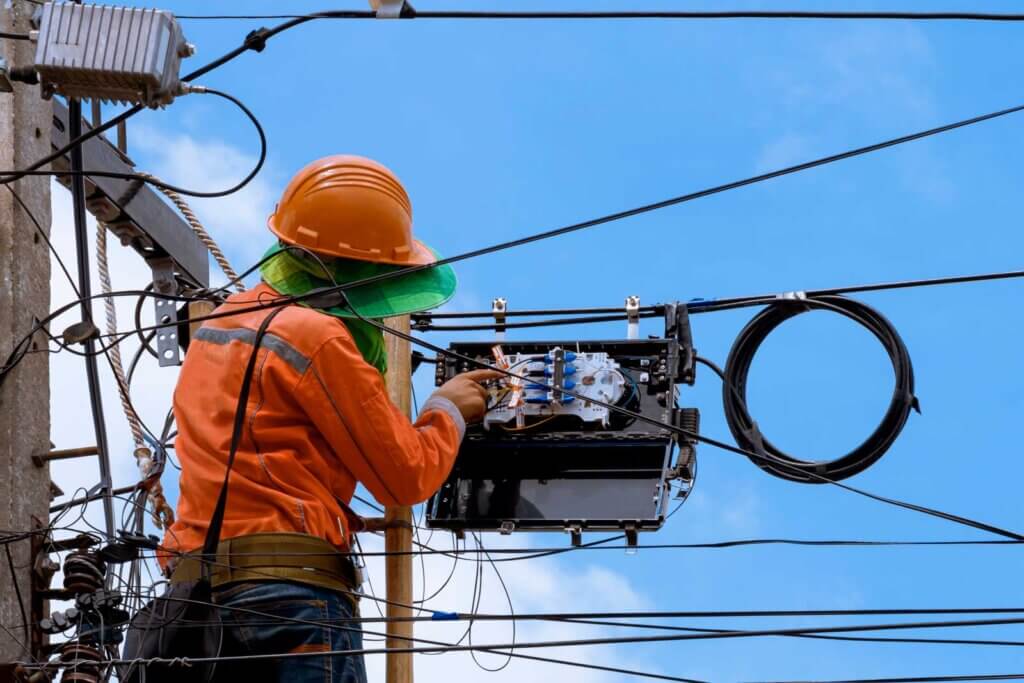 Rear view of technician on wooden ladder checking fiber optic cables in internet splitter box on electric pole against blue sky background